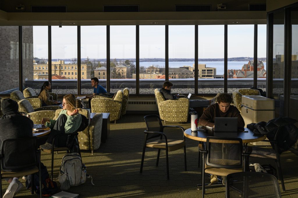Photo of students studying and chatting at round tables in the lounge of a high-rise building with windows looking out over Lake Mendota.