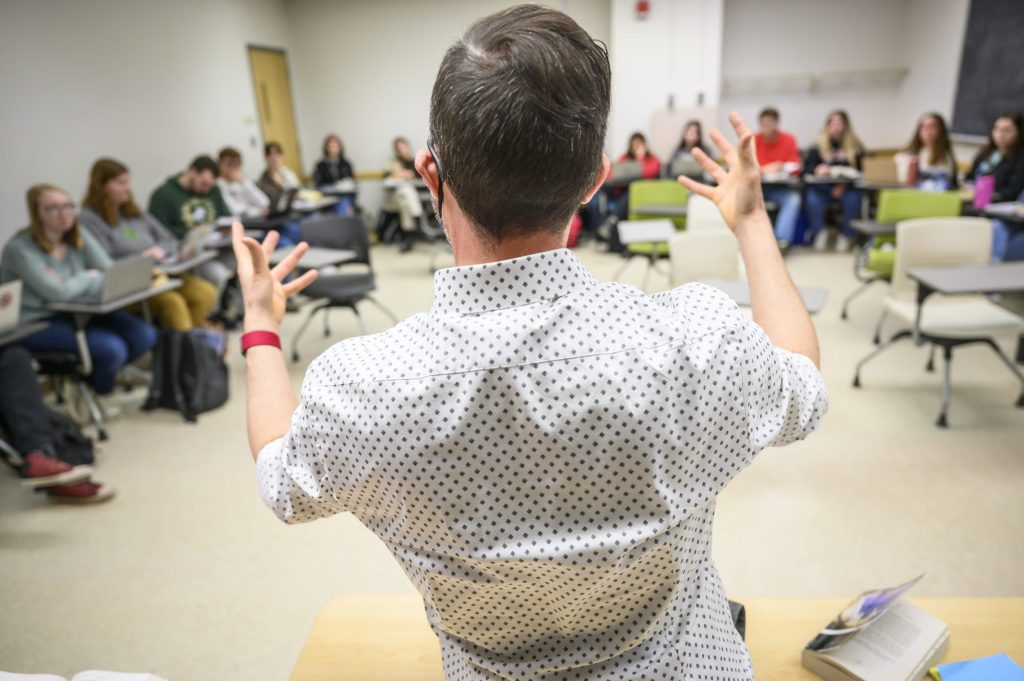 Photo of an instructor with his back to the photographer, facing a class of students seated in front of him and speaking to them while gesturing with his hands.