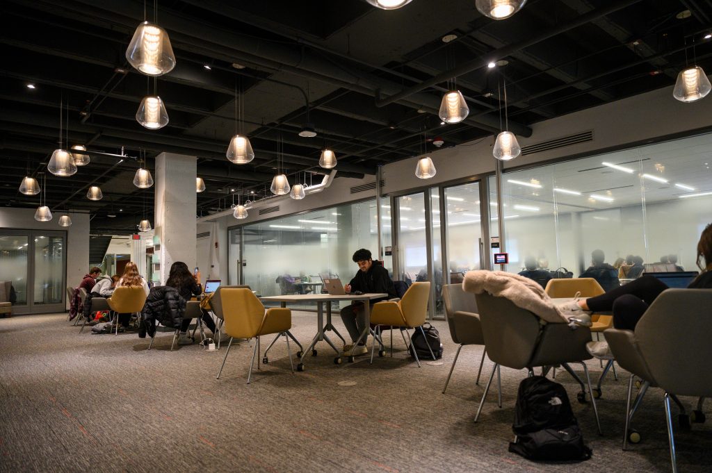 Photo of students studying at small round tables in a lounge in Grainger Hall with large windows behind them.